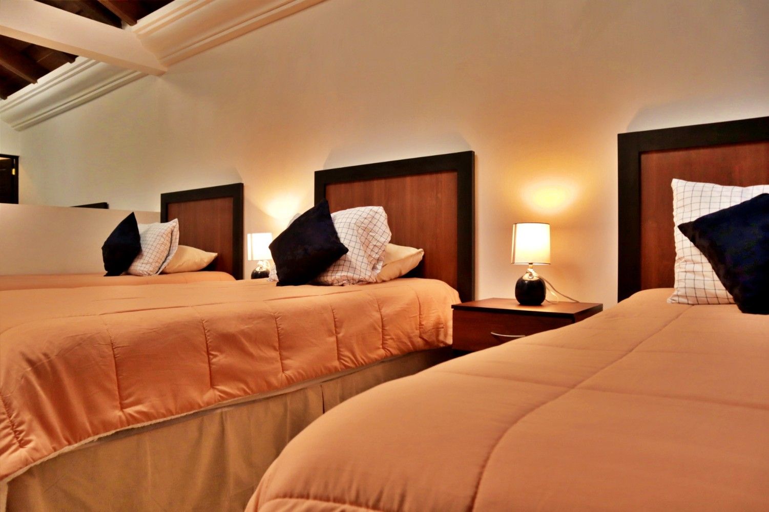 Tres camas bien hechas en una habitación de hotel con iluminación cálida, ropa de cama beige y lámparas de noche.