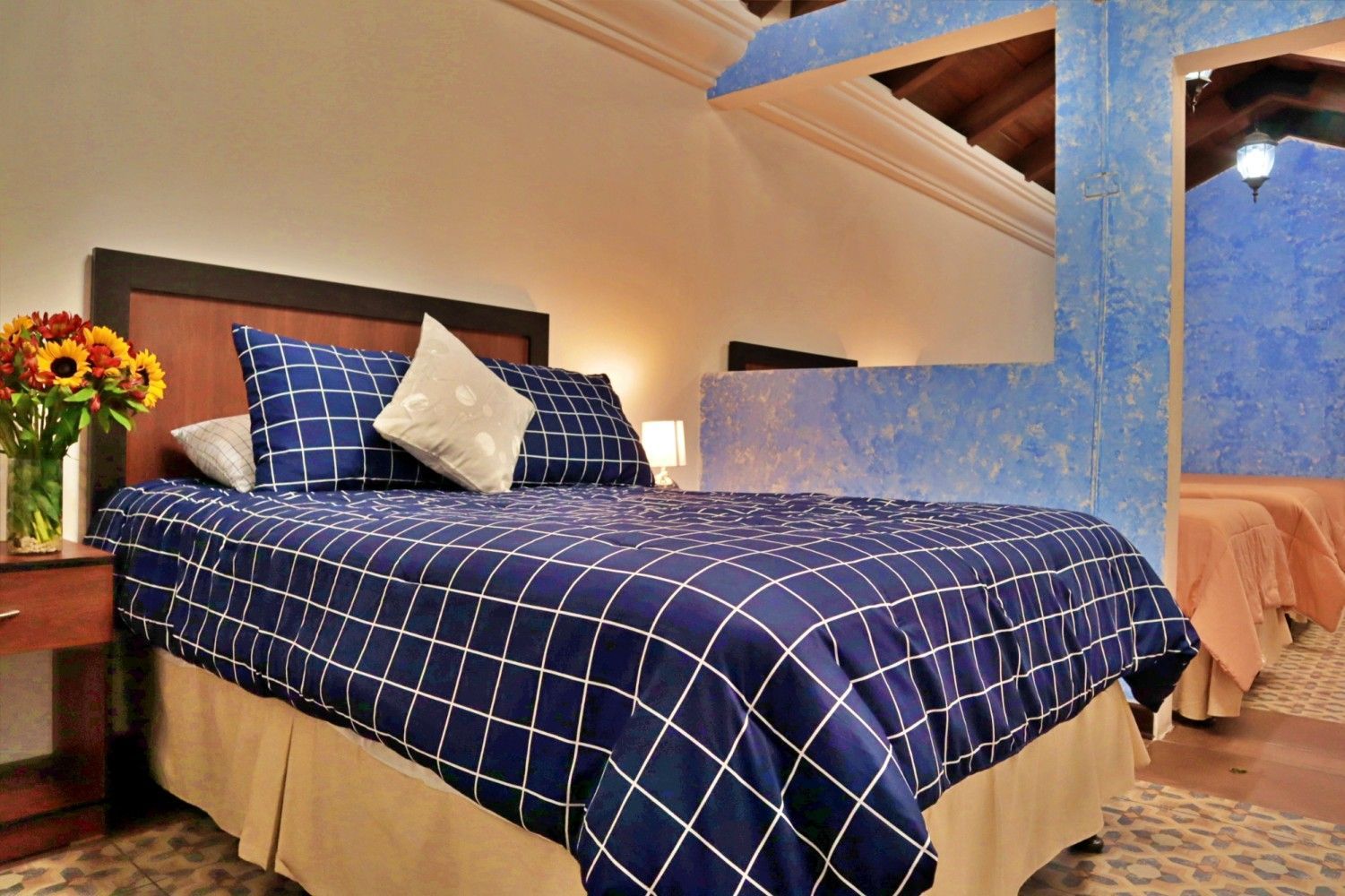 Habitación acogedora con una colcha a cuadros azules, almohadas y un jarrón de flores junto a la cama.