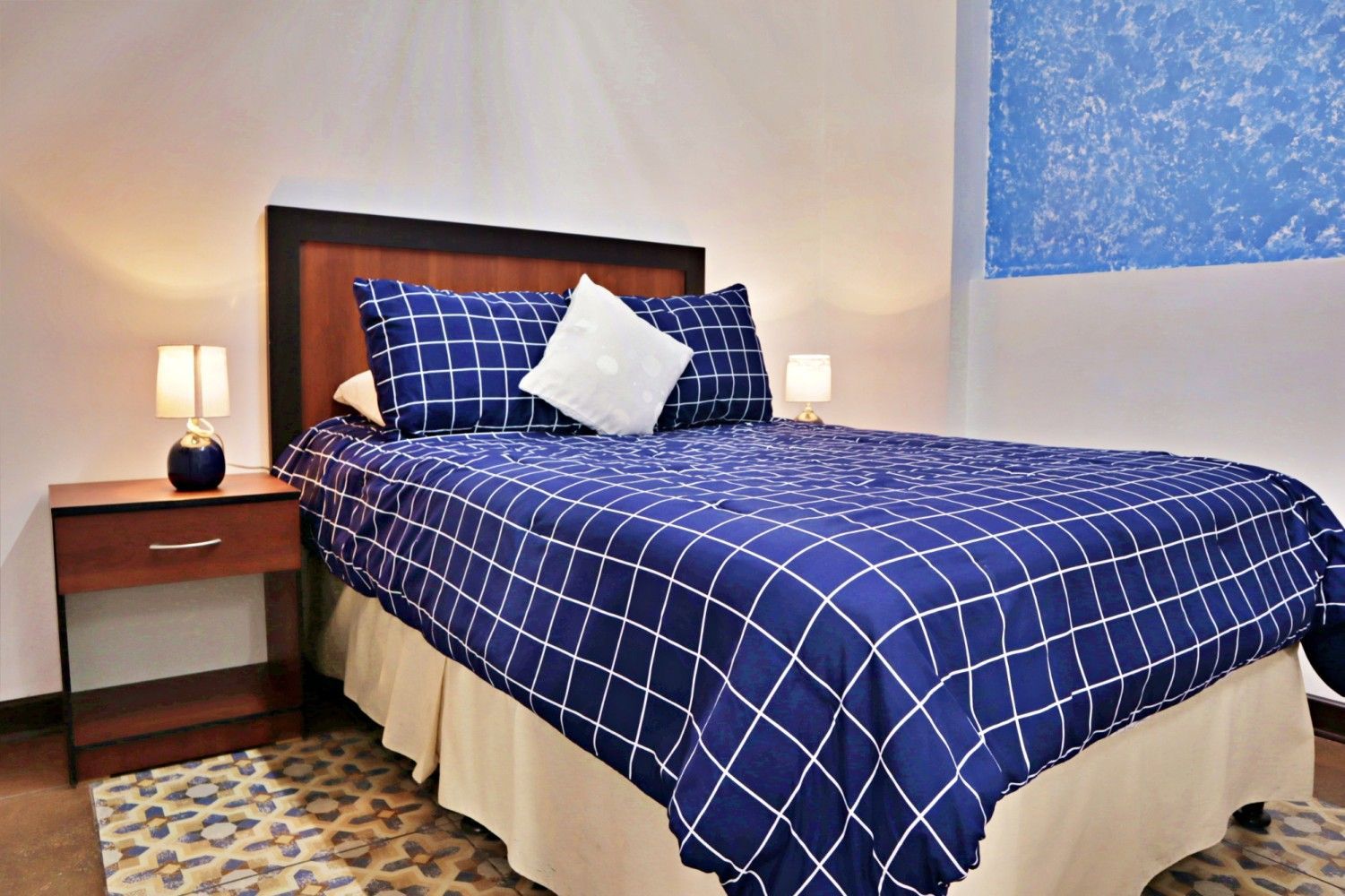 Dormitorio con una colcha de cuadros azules, almohada blanca, mesitas de noche y lámparas cálidas a cada lado.