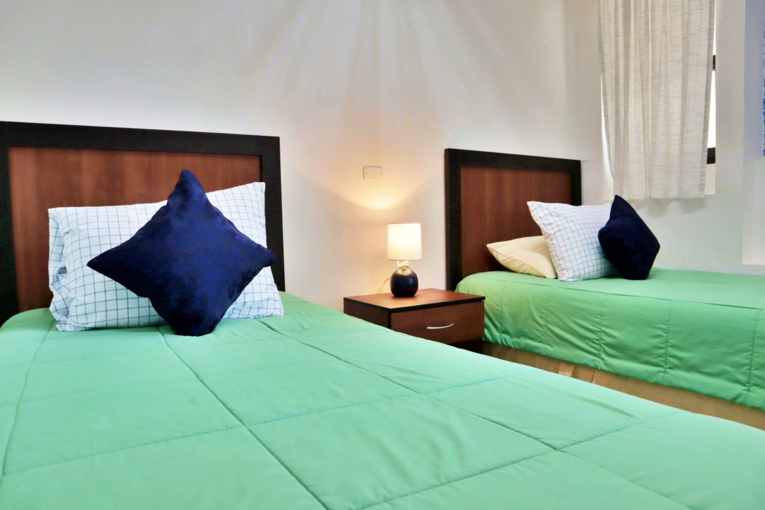 Dos camas bien hechas con sábanas verdes y almohadas azules en una luminosa habitación de hotel.