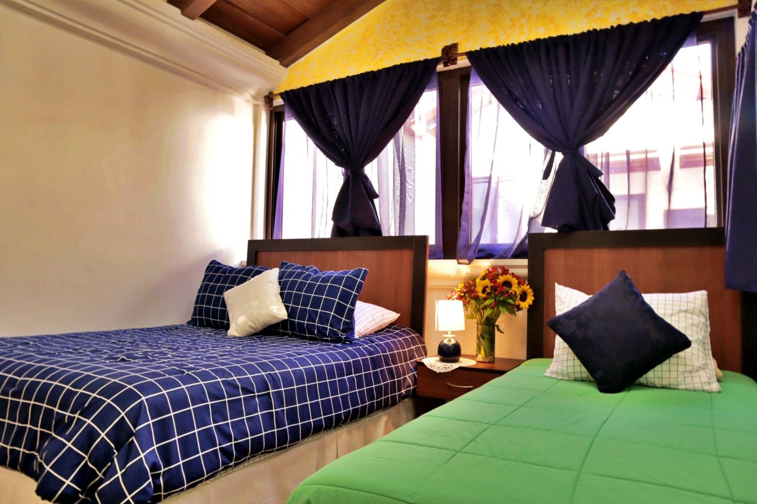 Habitación luminosa con dos camas, cortinas oscuras, cabecero de madera y un pequeño jarrón de flores entre ellas.