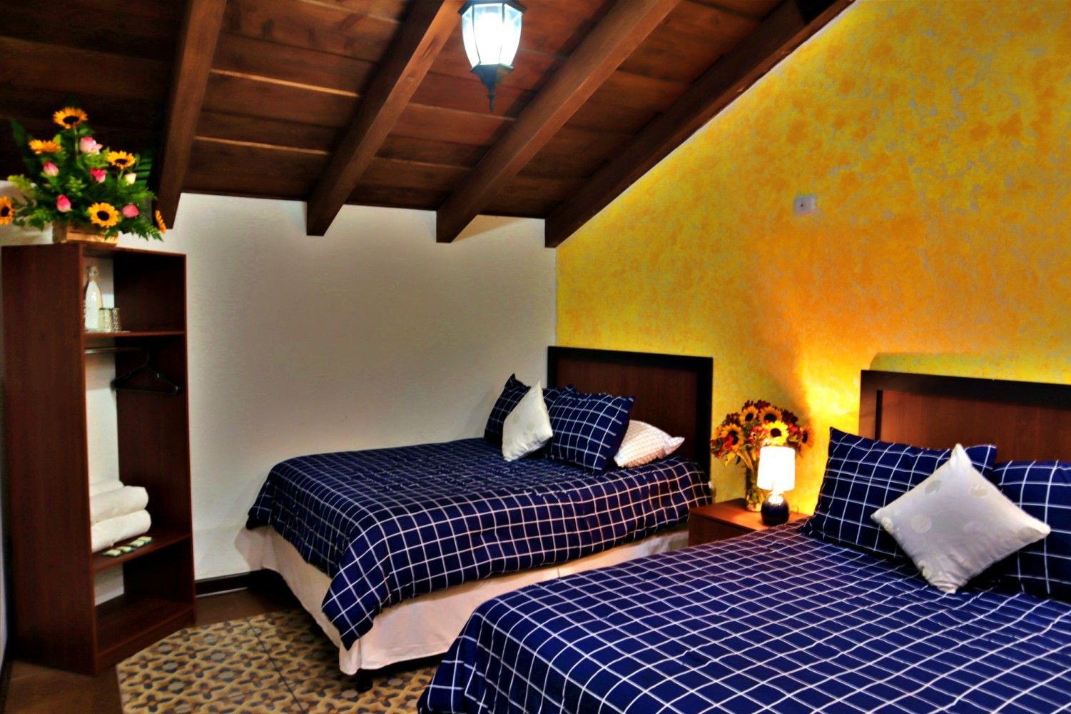 Acogedora habitación con dos camas de cuadros azules, paredes de un cálido color amarillo y techo con vigas de madera.