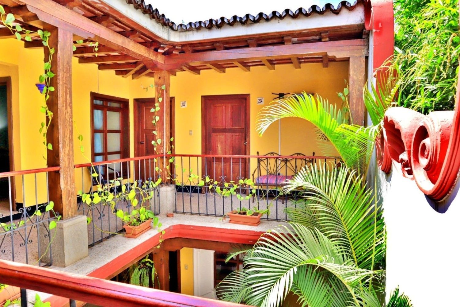 Patio soleado con paredes amarillas, puertas de madera, barandilla de balcón y exuberantes plantas tropicales.