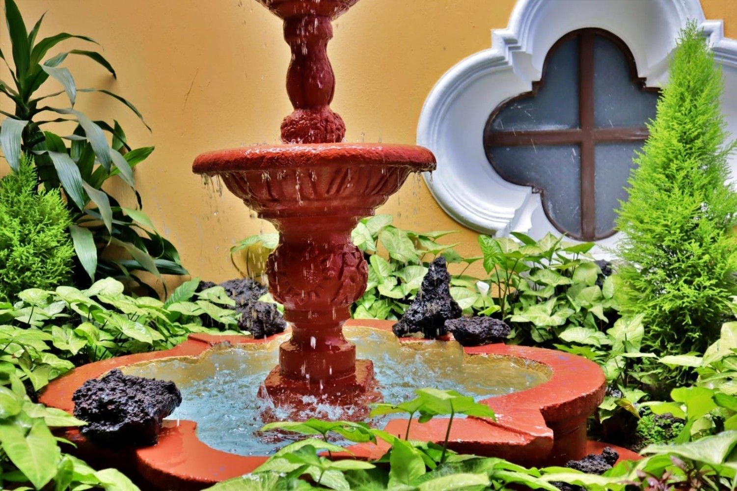 Fuente roja escalonada en un jardín exuberante junto a una ventana blanca con bordes festoneados y follaje verde.