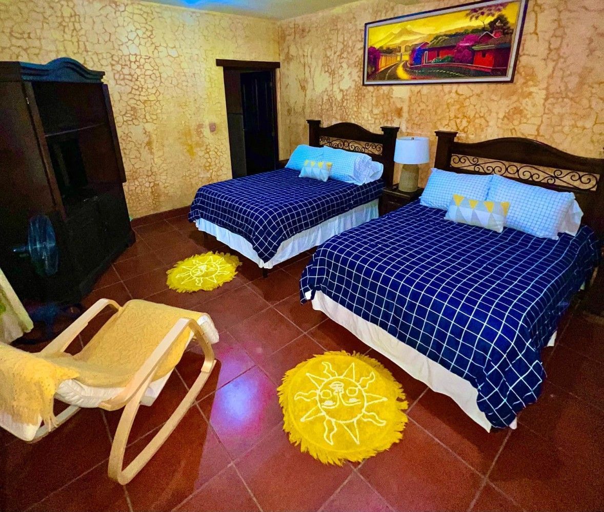Habitación acogedora con dos camas con fundas azules, paredes amarillas estampadas, suelo de baldosas rojas y una mecedora.