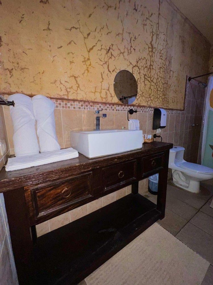 Mueble de baño con lavabo y toallas blancas, encimera de madera, espejo redondo e inodoro junto a una ventana.