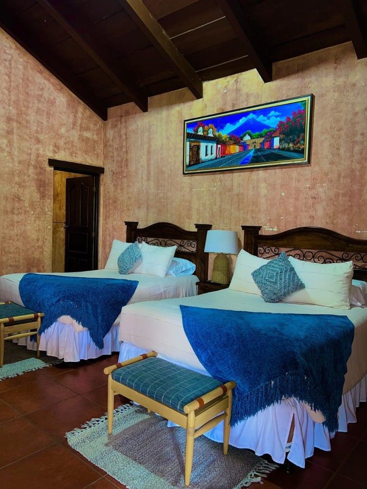 Habitación rústica con dos camas individuales, mantas azules, techo de madera y coloridos cuadros en las paredes.