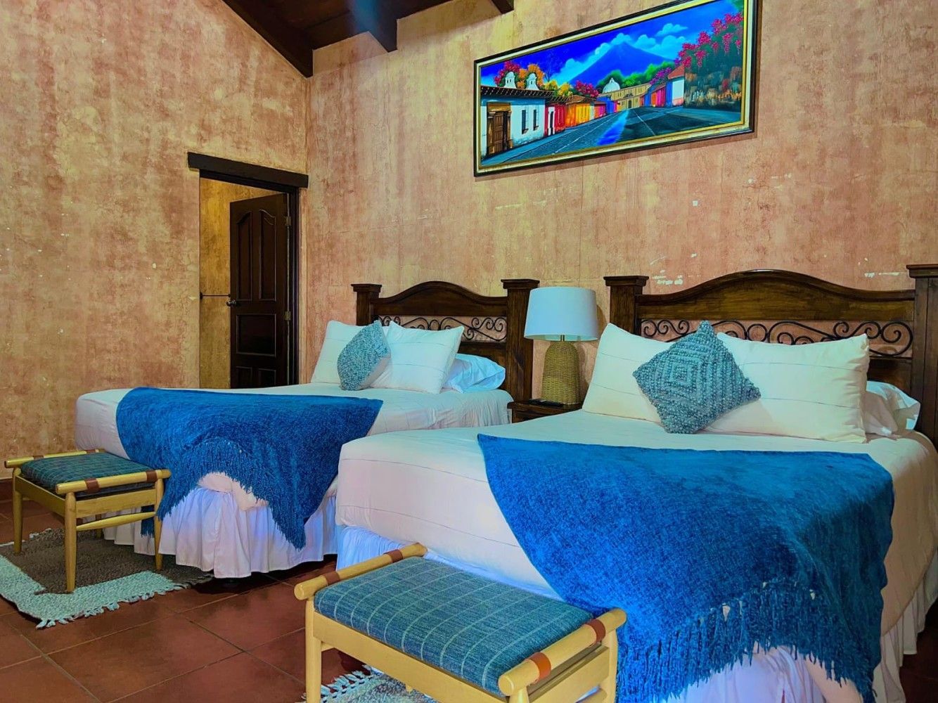 Dos camas individuales en una acogedora habitación con mantas azules, muebles de madera y un colorido cuadro en la pared.
