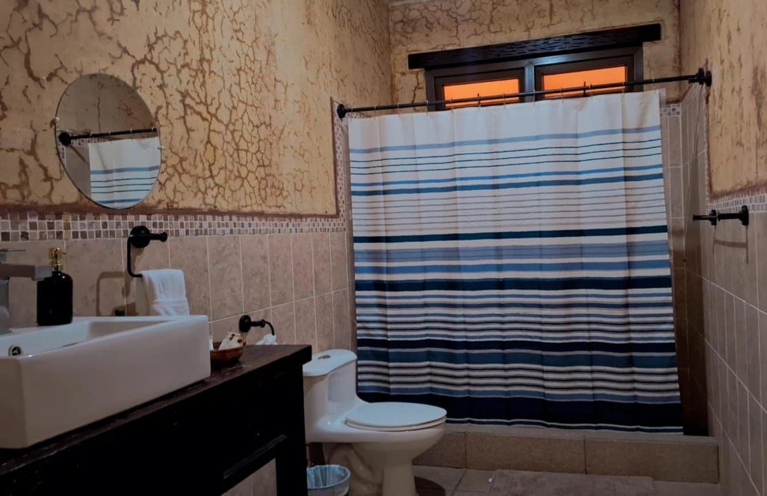 Baño con lavabo, inodoro, espejo y una cortina de ducha a rayas debajo de una pequeña ventana.