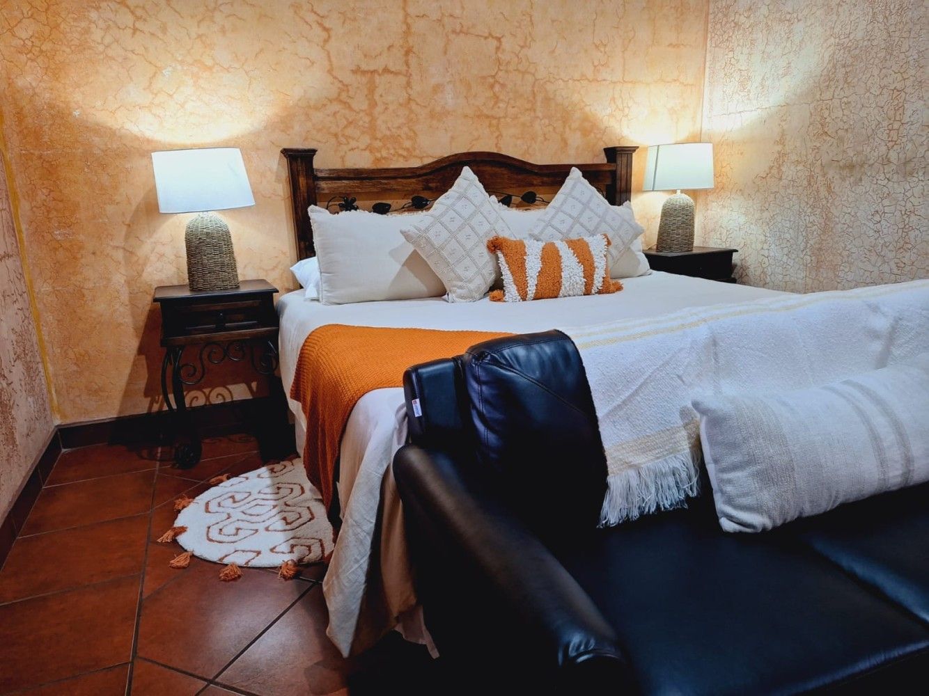 Habitación acogedora con cama de madera, detalles en naranja, lámparas de noche y un sillón de cuero frente a ella.