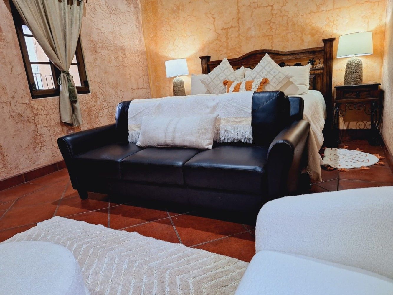 Habitación de hotel con sofá de cuero negro a los pies de la cama, iluminación cálida y suelo de baldosas rojas.