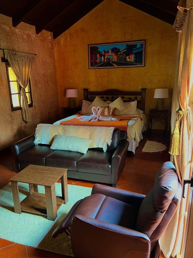 Dormitorio rústico con cama, sillas de cuero, mesa de centro y paredes de color amarillo cálido.