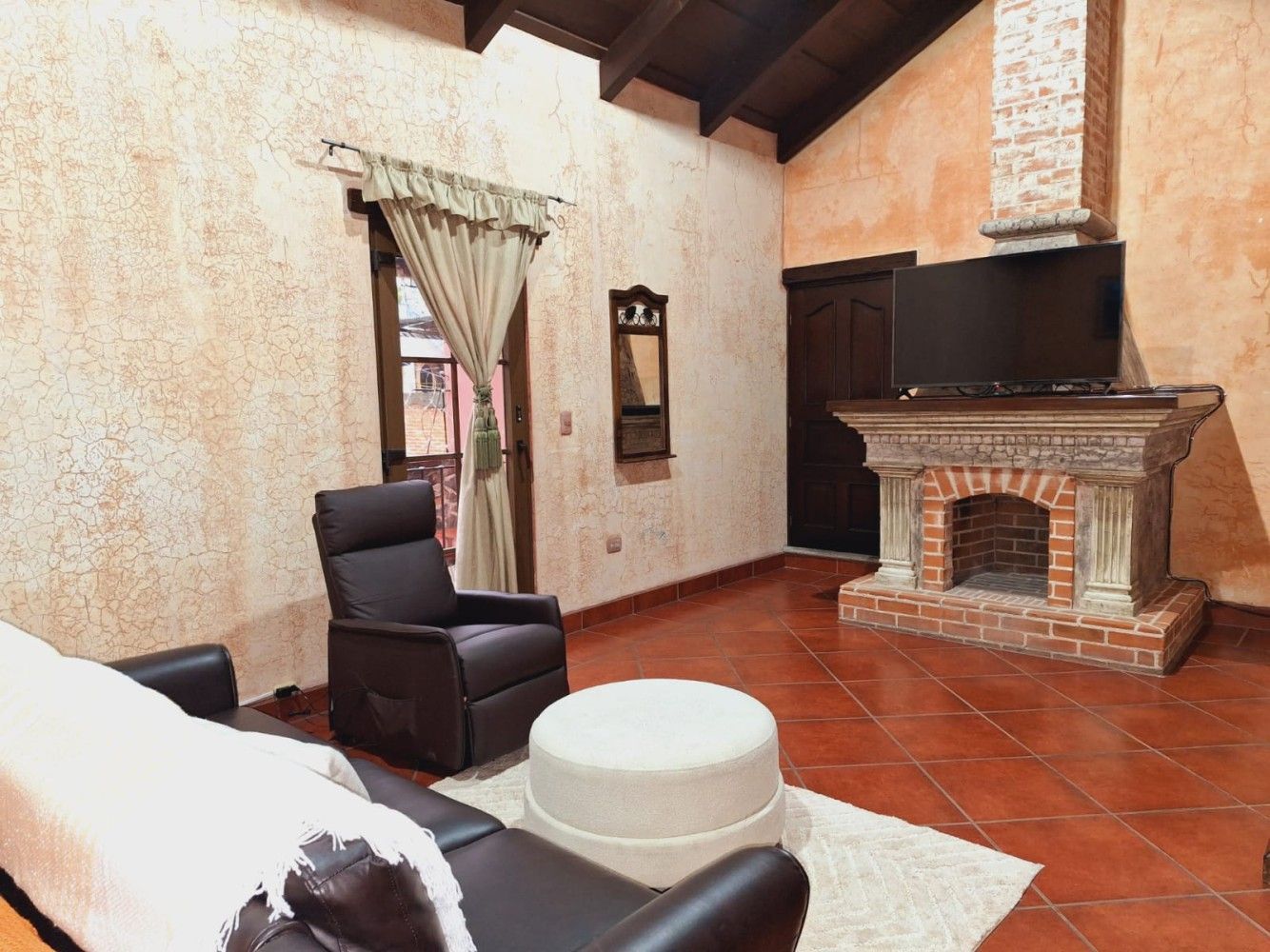 Sala de estar rústica con sillones de cuero, chimenea, suelo de baldosas y ventana con cortinas.
