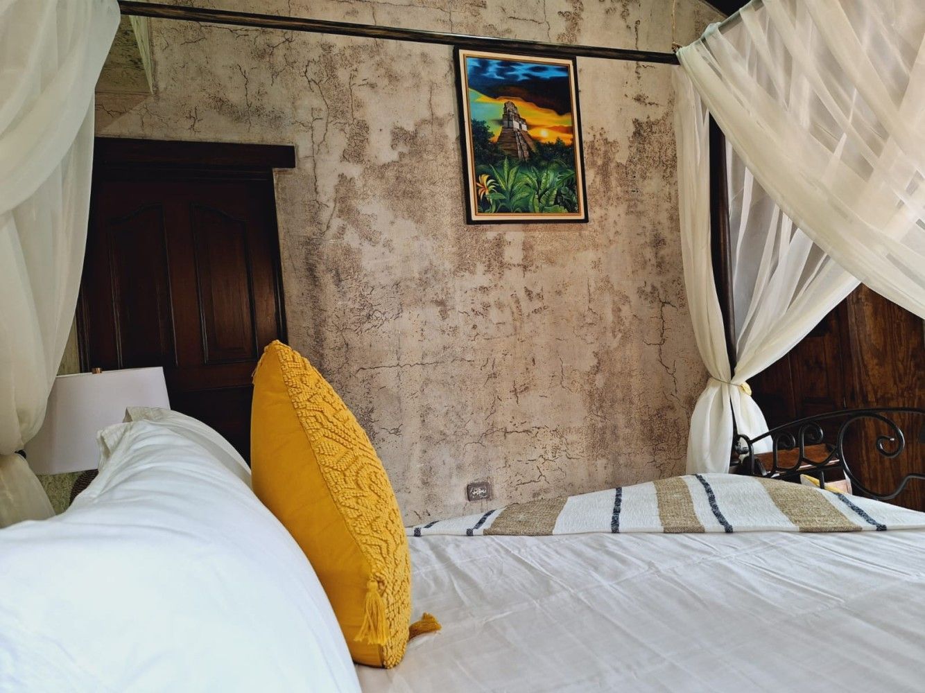 Dormitorio rústico con cama blanca, almohada amarilla, cortinas transparentes y un cuadro enmarcado en la pared.