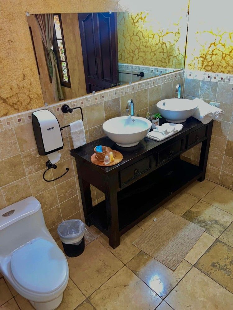Baño con tocador de doble lavabo, espejo, inodoro y suelo de baldosas beige.