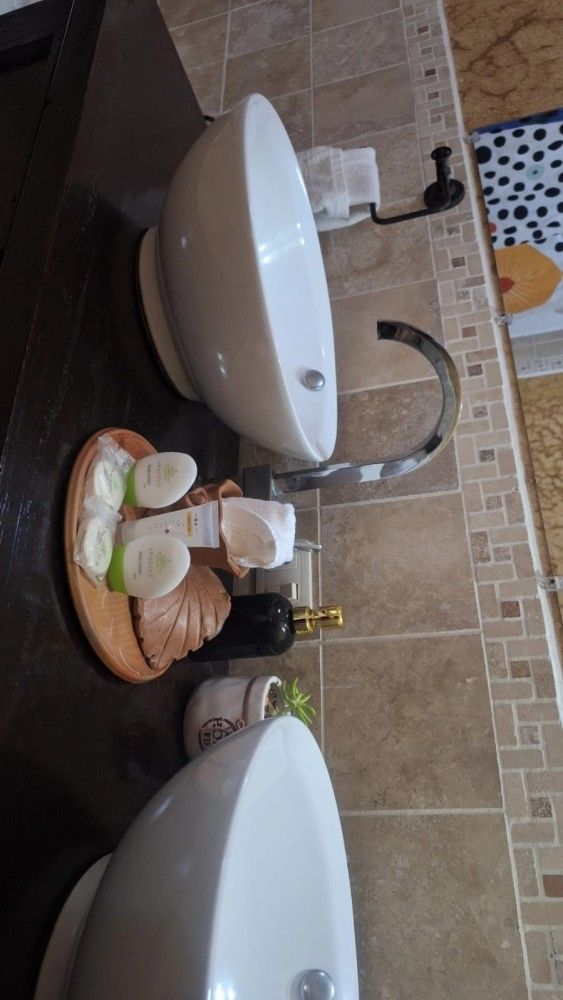 Encimera de baño con dos lavabos blancos, jabón, cepillos de dientes y toallas apiladas bajo un grifo cromado.