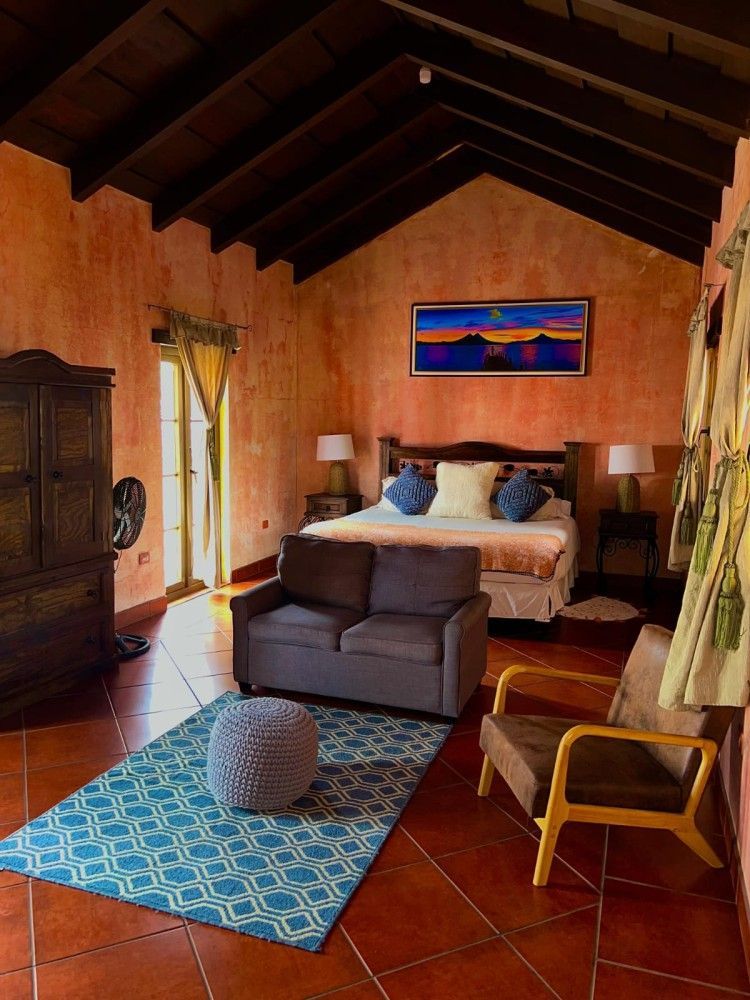 Acogedora habitación con techo de madera, cama, alfombra azul, sillón y coloridos cuadros en la pared.
