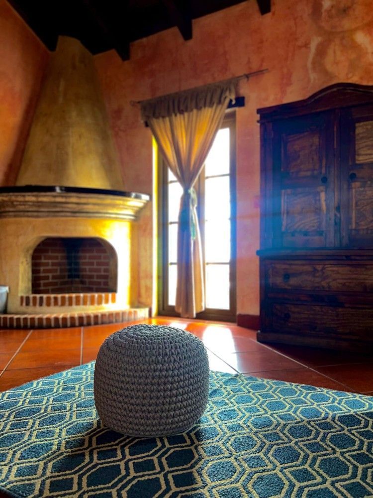 Habitación acogedora con chimenea, ventanal alto, alfombra azul y otomana tejida sobre baldosas de terracota.
