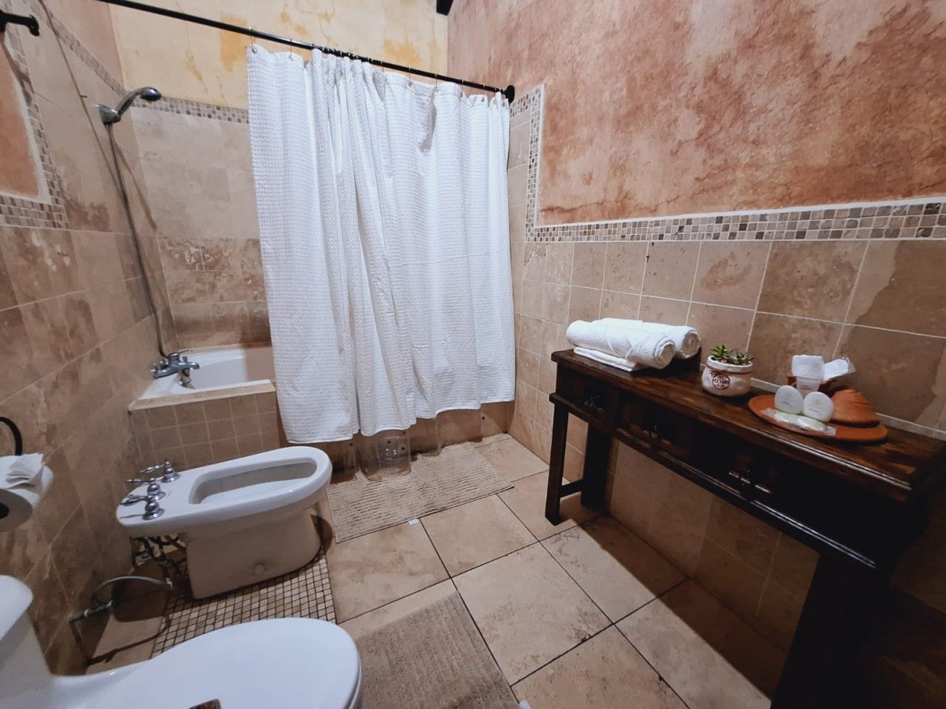 Baño pequeño con azulejos, cortina blanca, inodoro, lavabo y tocador oscuro con artículos de aseo y toallas.