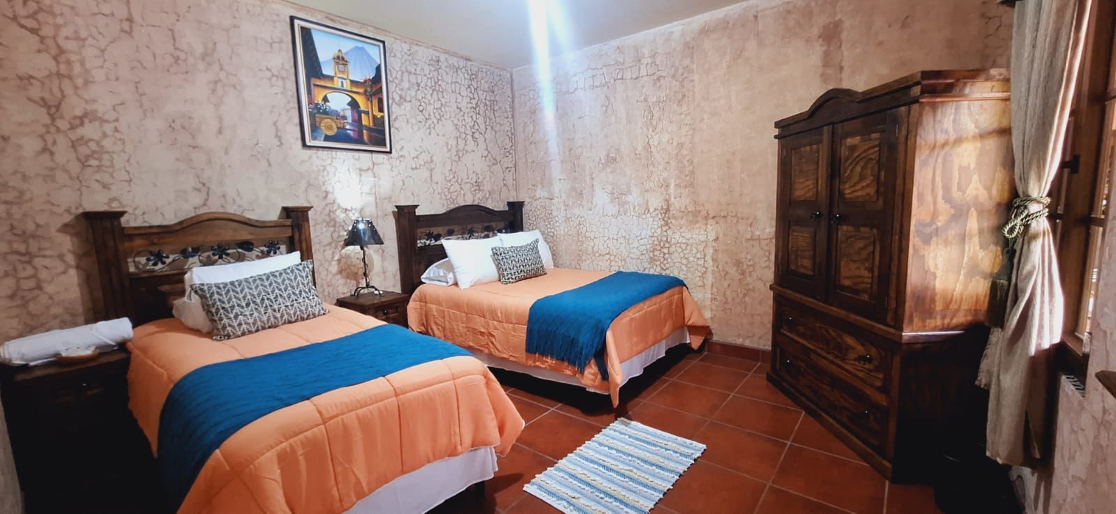 Habitación pequeña con dos camas individuales, ropa de cama azul anaranjado, armario de madera y suelo de baldosas.