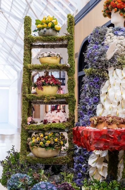 A moss-covered ladder displays bowls of flowers and fruit, standing beside a mannequin decorated with floral arrangements.