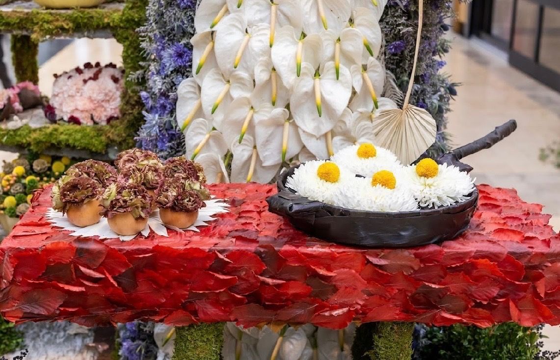 A red floral display featuring white calla lilies, a skillet of white daisy-like flowers, and small clusters of brown buds.