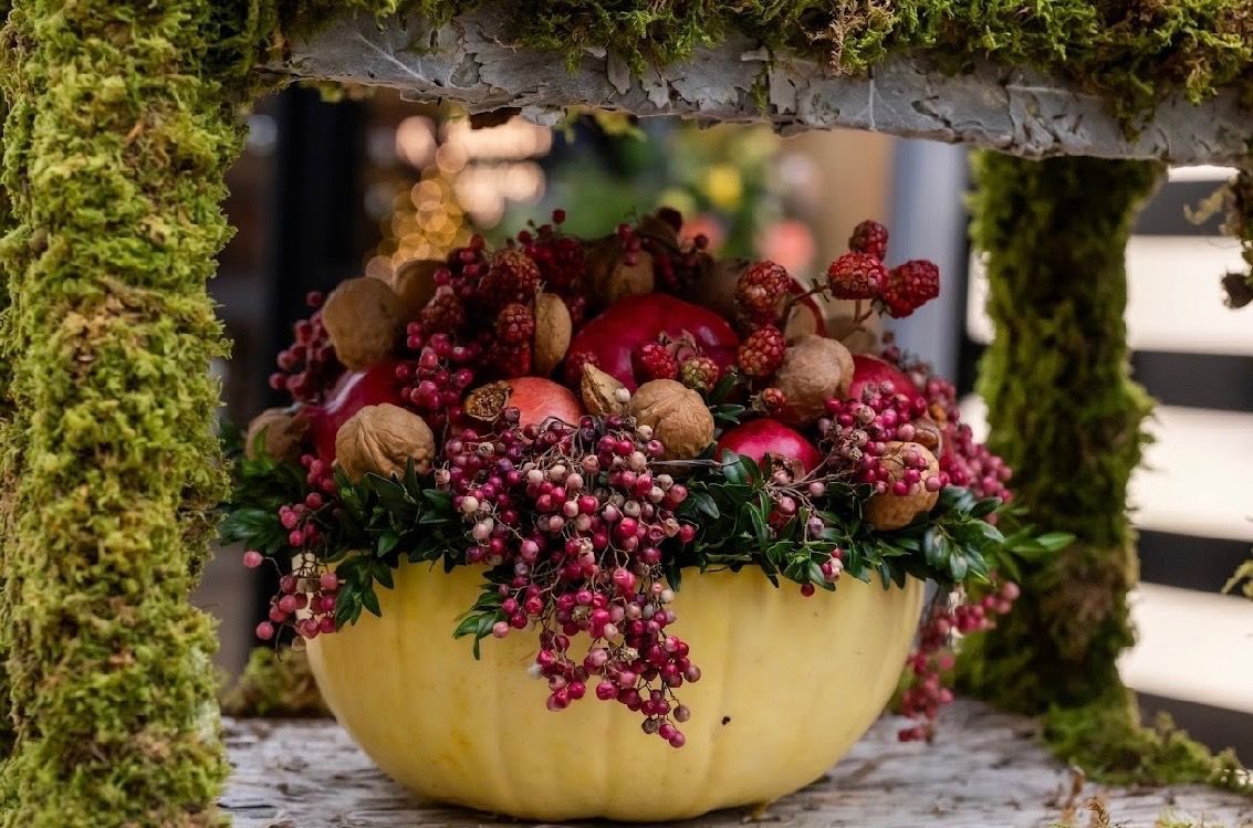 A centerpiece in a light yellow bowl filled with red berries, walnuts, and apples, framed by moss-covered wooden structures.