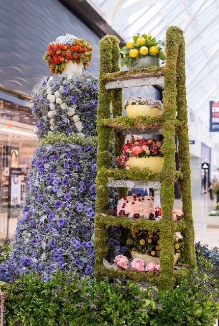A mannequin dressed in purple floral attire stands beside a moss-covered ladder displaying bowls of colorful flowers.