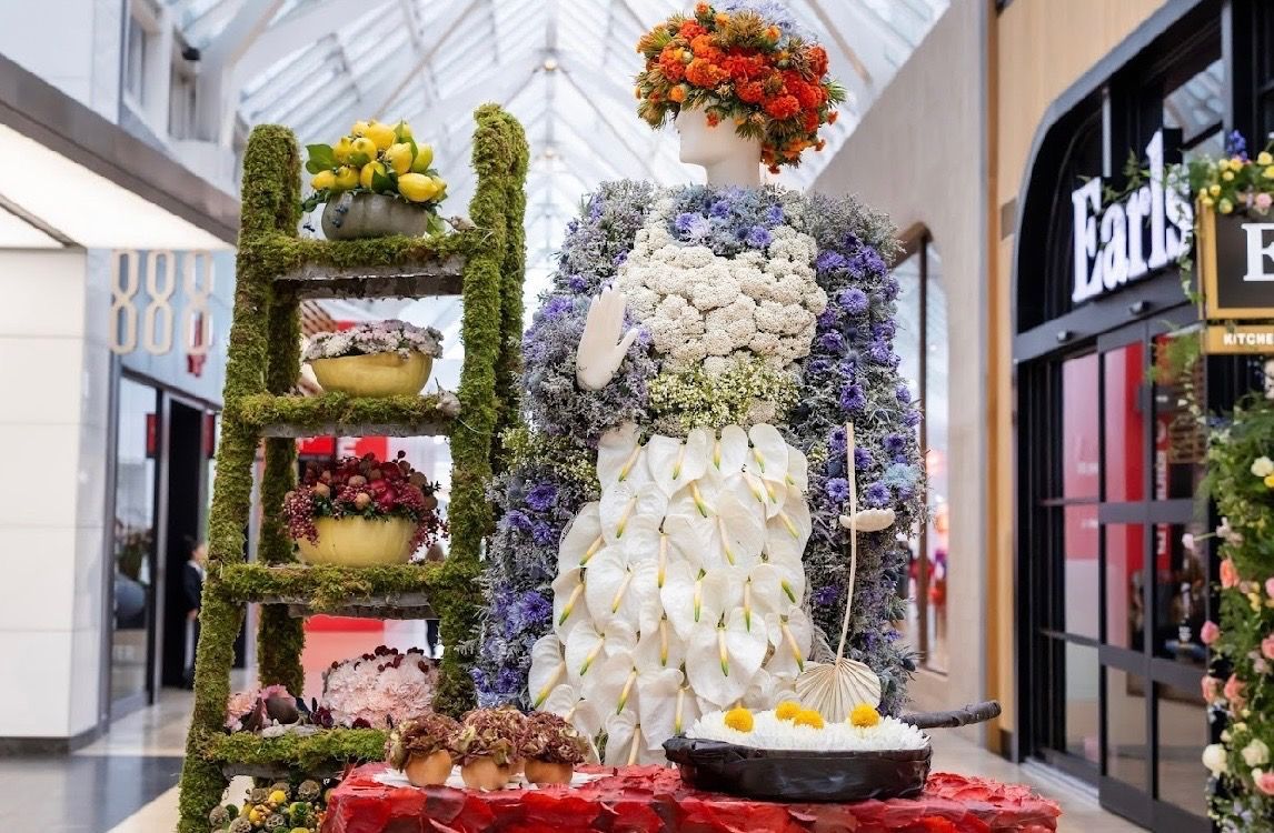 A floral display featuring a mannequin wearing a dress made of white and blue flowers, next to a moss-covered shelf.