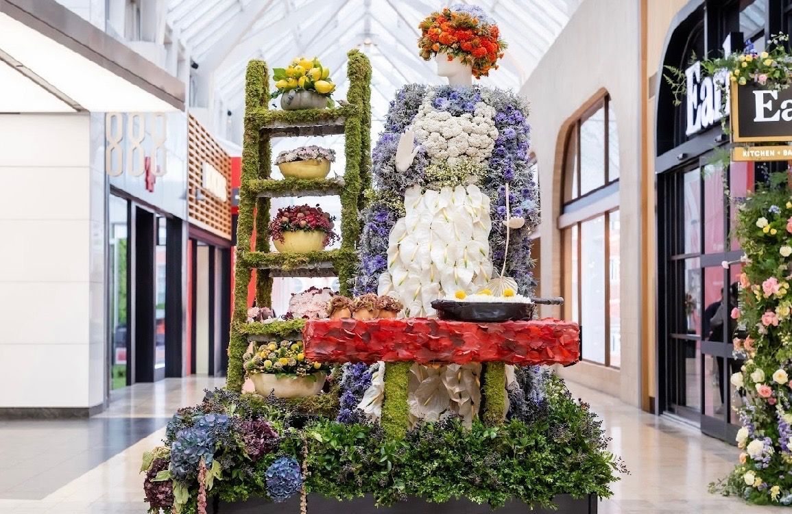 A floral display in a shopping mall featuring a mannequin dressed in flowers standing behind a table with a skillet.
