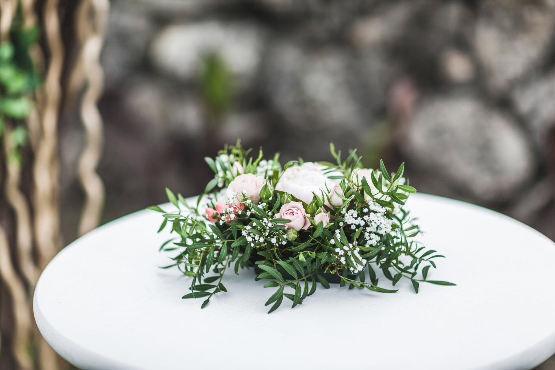 A small floral centerpiece of white and pale pink roses with green foliage sits on a round white table outdoors.