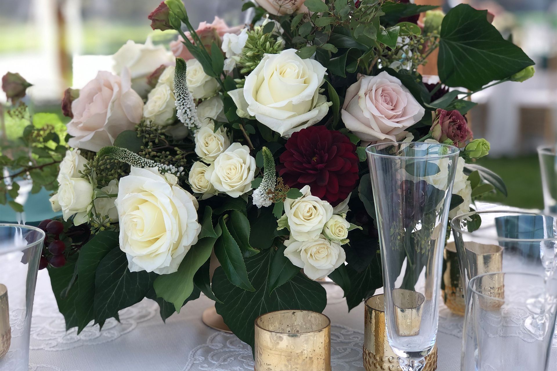 A floral centerpiece with cream, blush, and dark red roses surrounded by greenery, set on a table with gold votives.