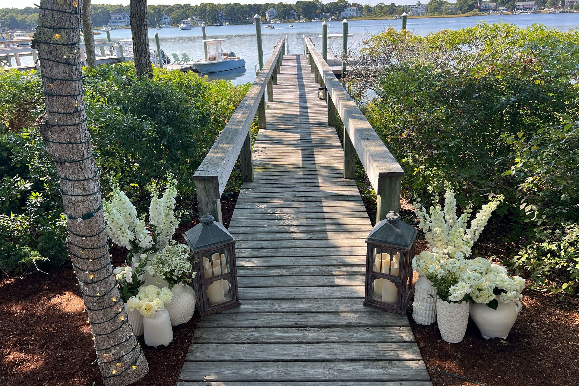 A wooden dock extends over water, flanked by lanterns and white floral arrangements at the entrance.