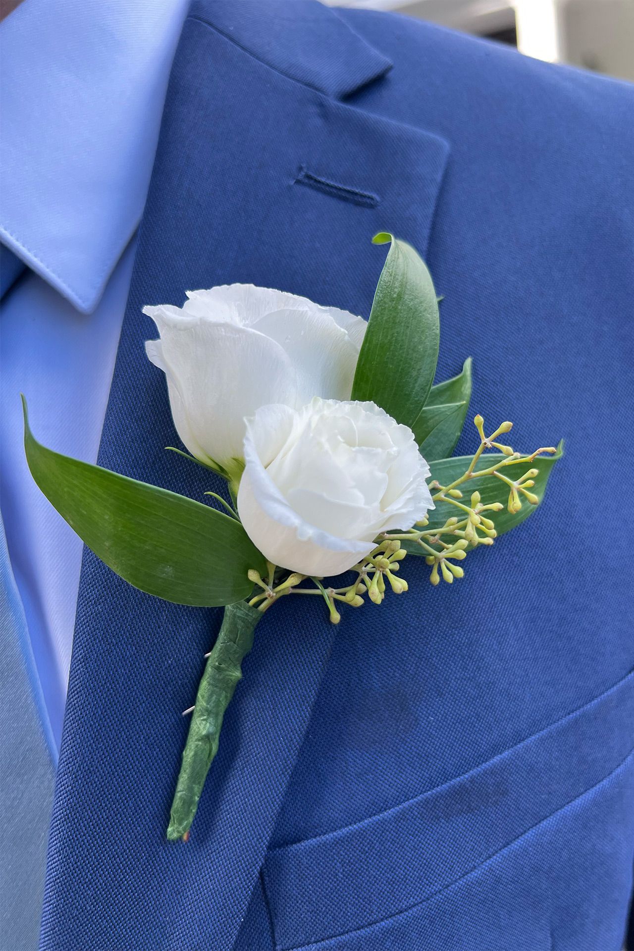 A white rose boutonniere with green leaves and small yellow buds pinned to a blue suit jacket.