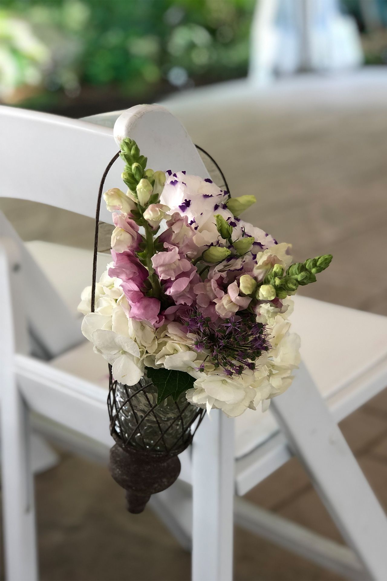 A metal hanging vase filled with white and pink wedding flowers attached to a white chair.