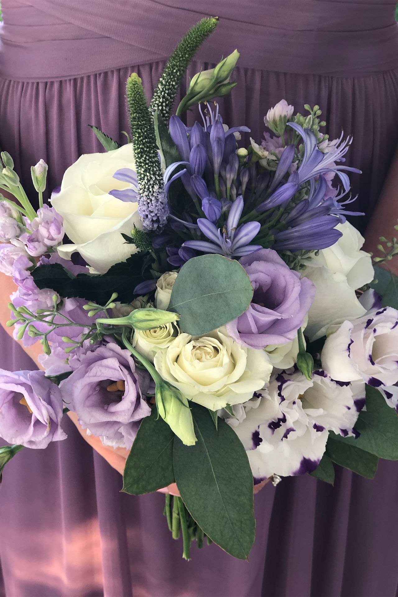 A bridesmaid in a purple dress holds a bouquet of white roses, lavender lisianthus, blue agapanthus, and green foliage.