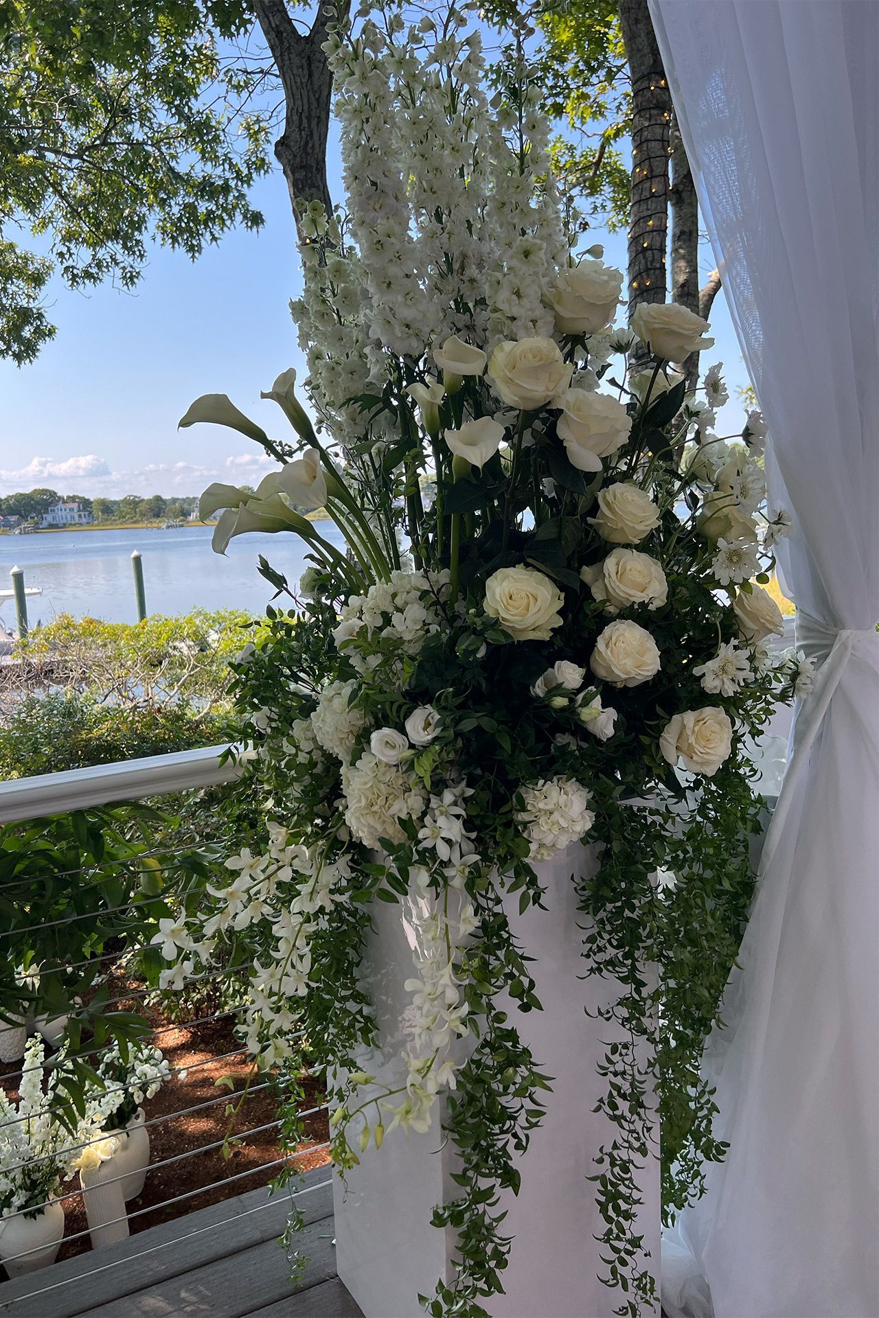 A large, elegant white floral arrangement sits on a pillar stand against a backdrop of a sunny waterfront.