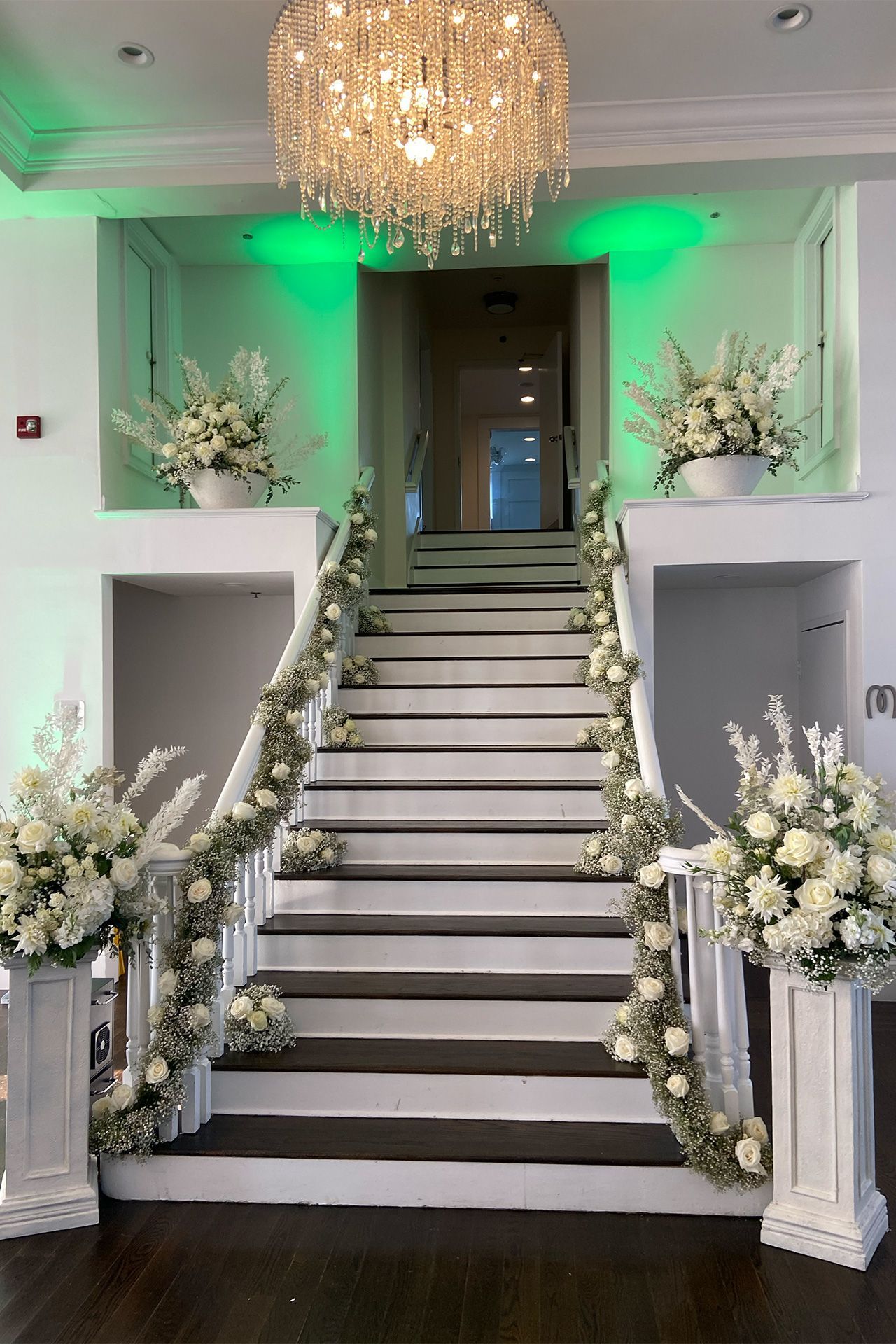 A staircase decorated with white floral arrangements and greenery, illuminated by a crystal chandelier and green uplights.