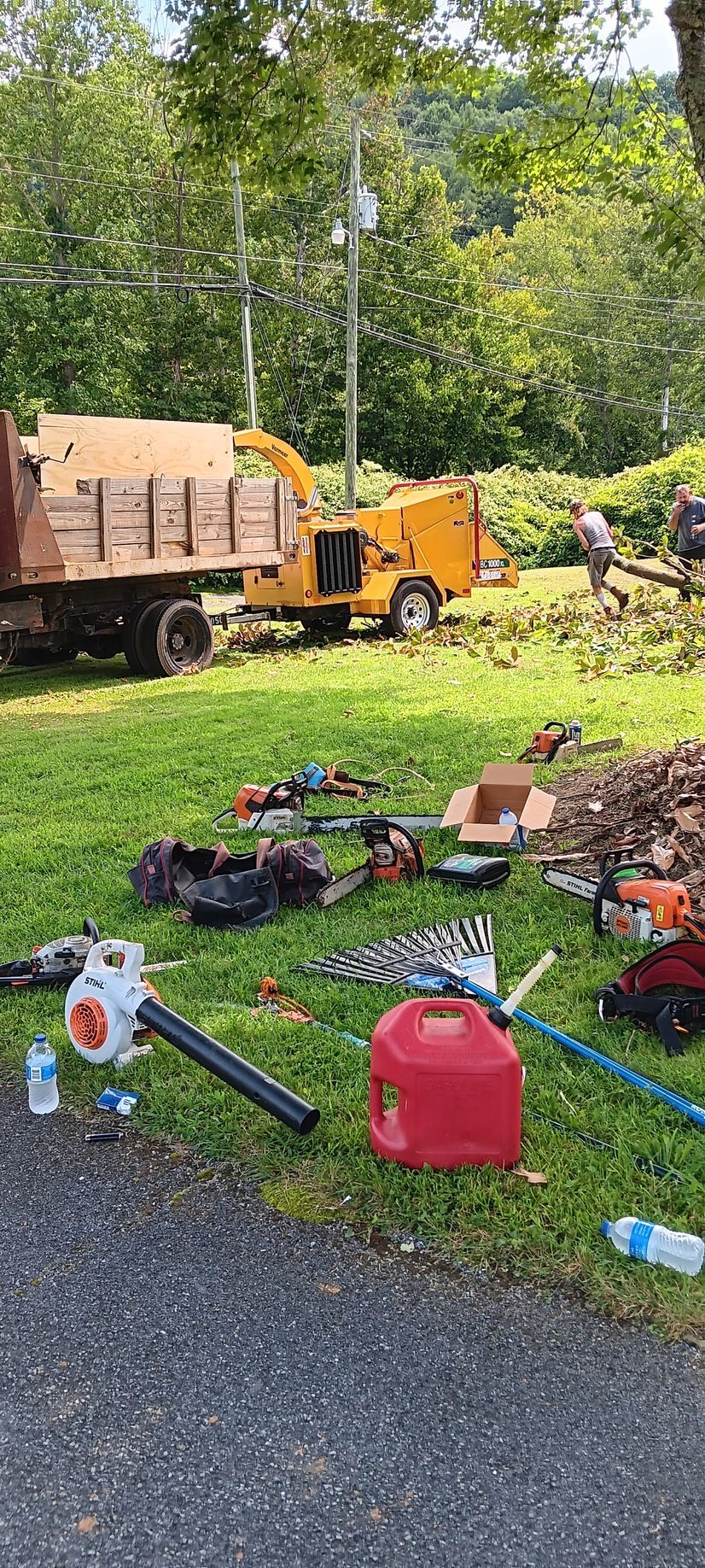 Yellow wood chipper beside a truck in a grassy yard, with tools and debris scattered in the foreground.
