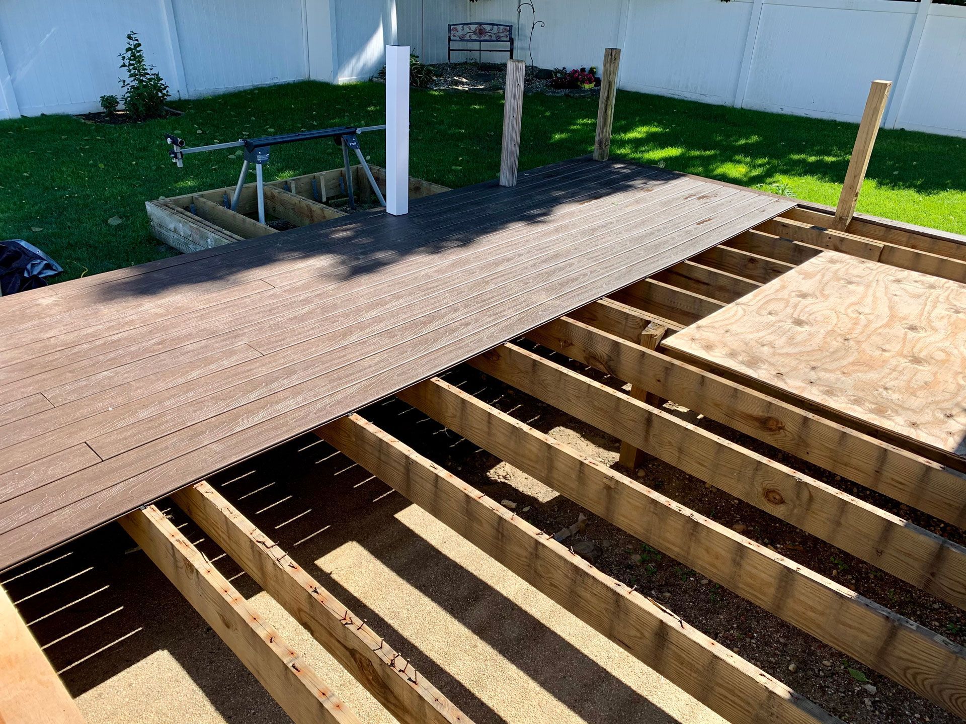 Partially built wooden deck framing beside a yard, with exposed joists and a finished section in sunlight