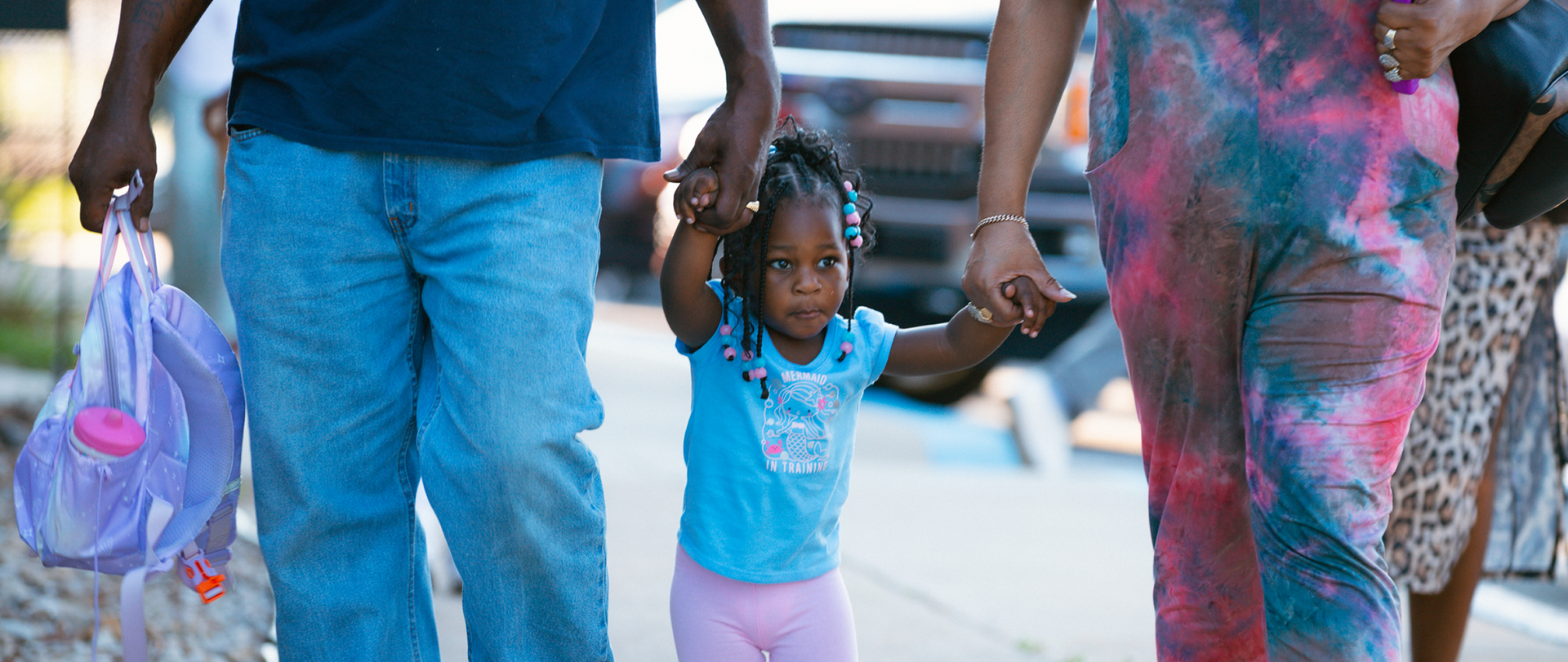 Two adults holding a toddler’s hands while walking outdoors, with plastic bags and colorful clothing.