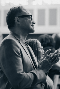 Man in glasses and blazer clapping at an indoor event, shown in side profile.