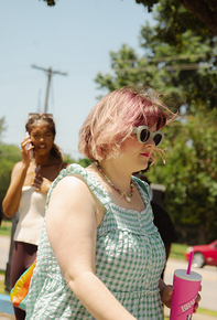 Person with pink hair and sunglasses walking outdoors, holding a pink drink bottle.