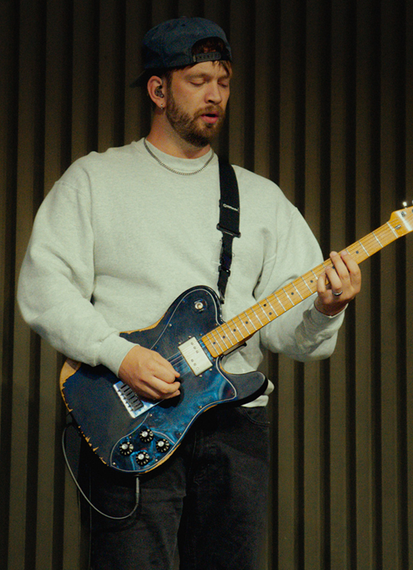 Musician playing a blue electric guitar onstage, wearing a white sweater and black cap.