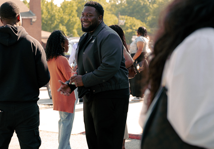 People standing outdoors in a sunny public space, with one man smiling toward the camera.