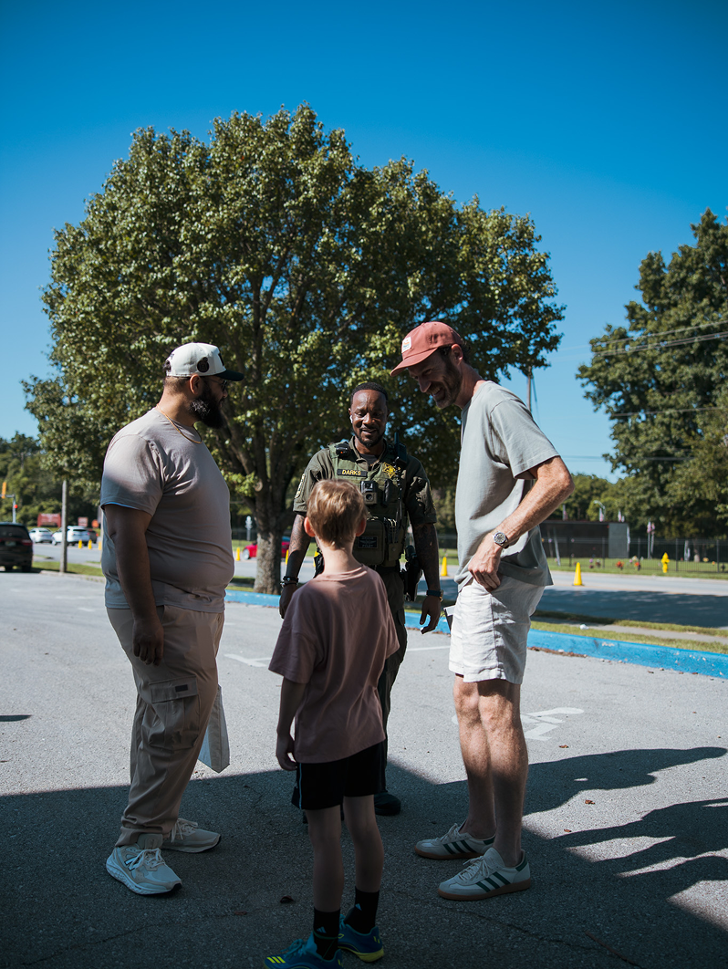 Three people standing together outdoors near a tree on a sunny day, talking on a paved path.