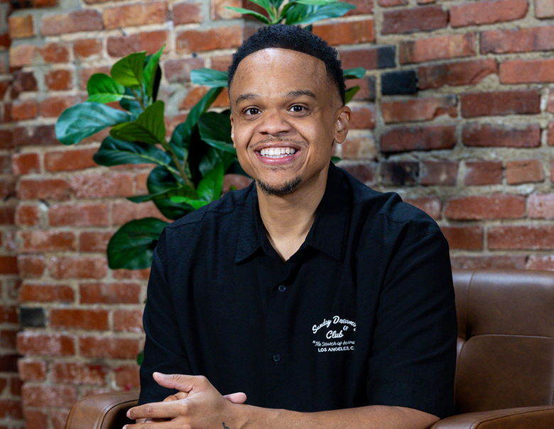 Smiling person in a black shirt seated indoors against a brick wall with a green plant behind them