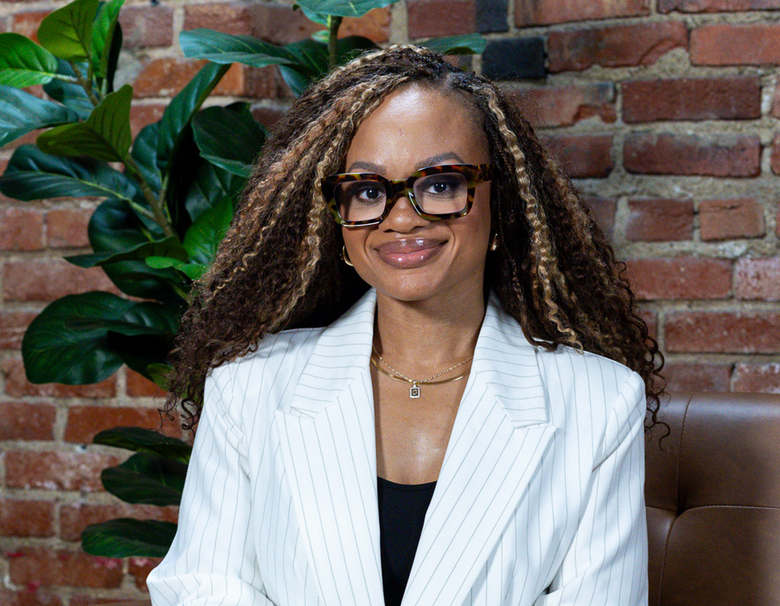 Woman in glasses and white blazer smiling against a brick wall with green plants.