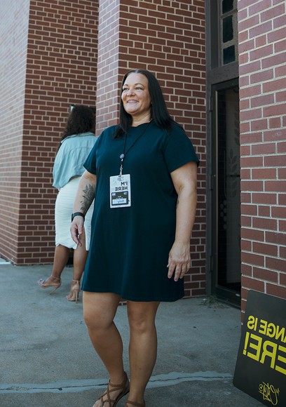 Woman in a dark dress and lanyard standing by a brick building, smiling outdoors.