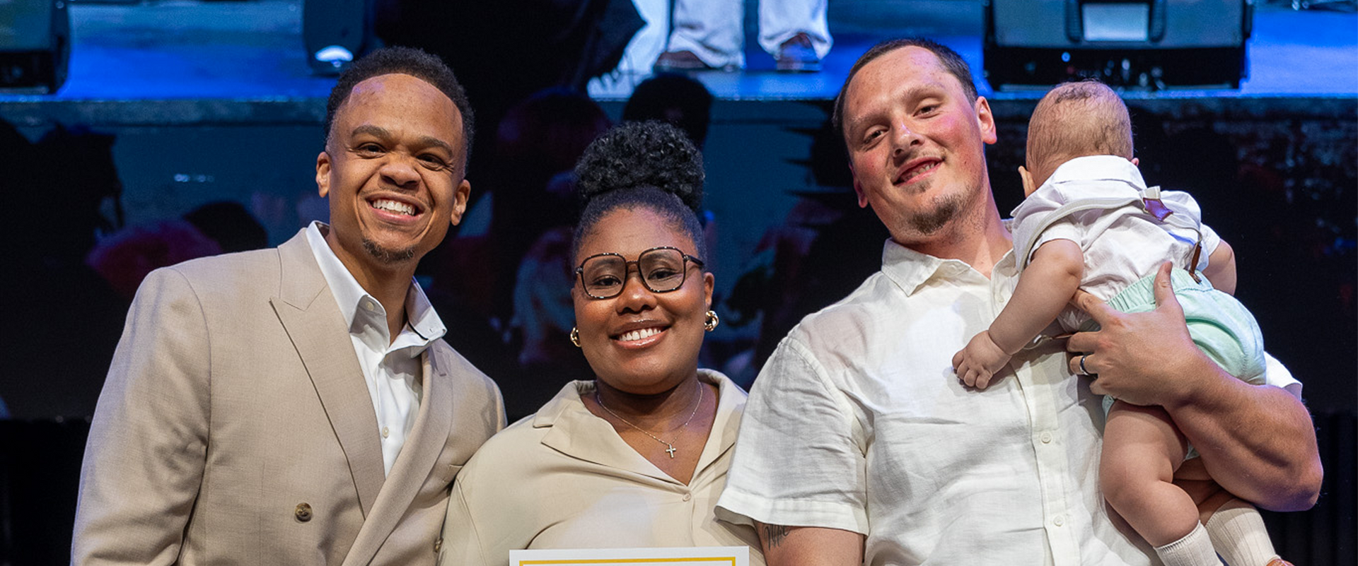 Three people smiling onstage, with one holding a baby and a blue-lit background